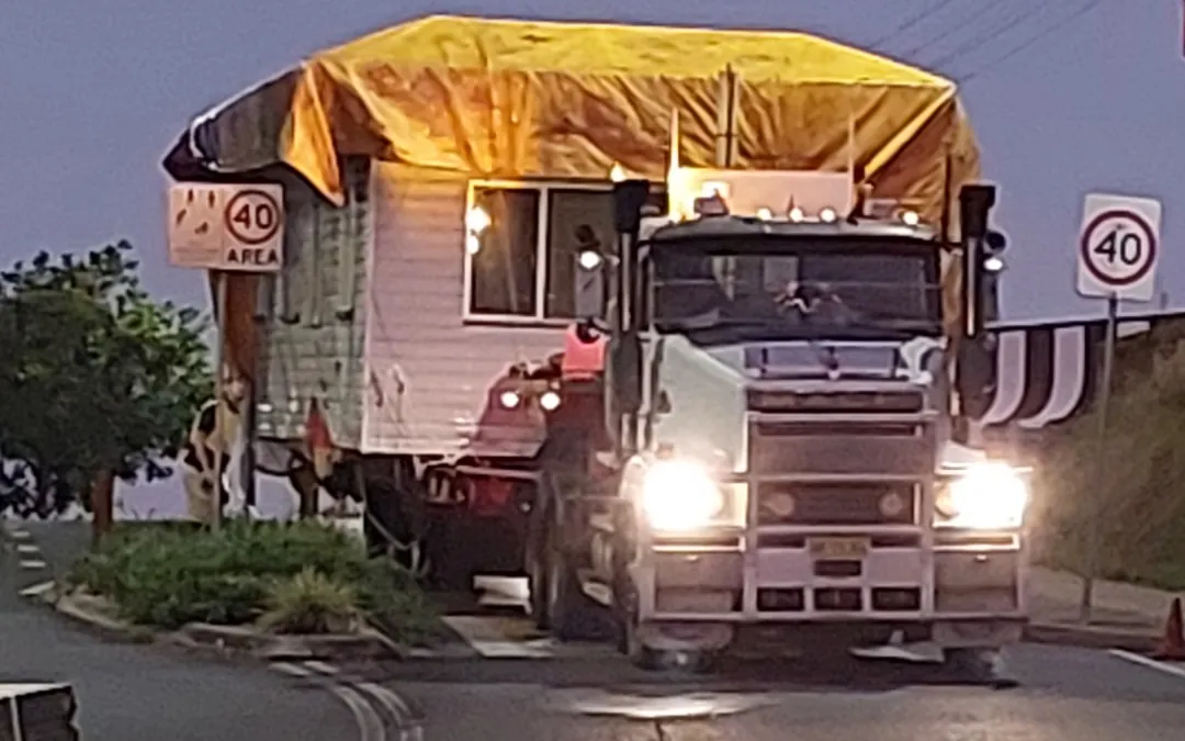 House under a tarp being relocated by truck through town at dusk